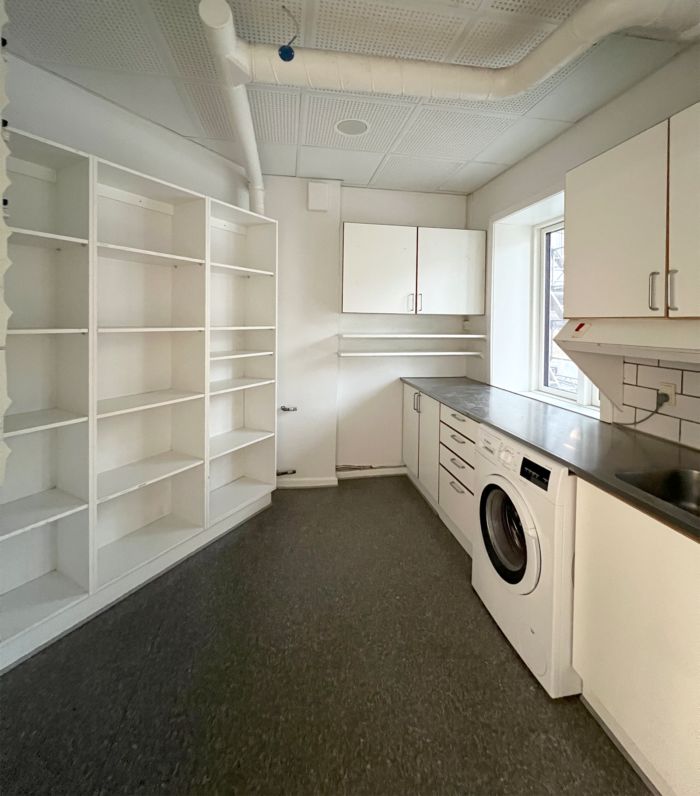 Hvid vaskeri med hvid vaskemaskine, skraner og et lager med stel, hvor du kan opbevare købsvarer. Hvidt loft og vægge.