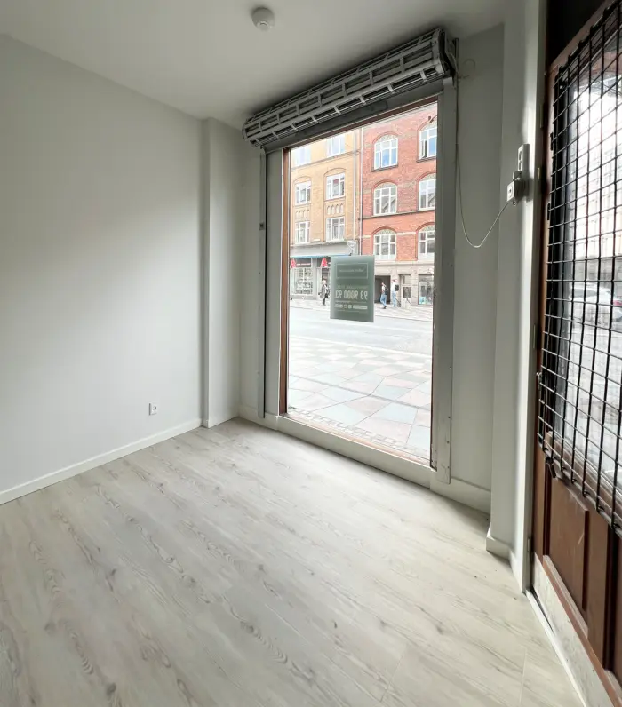 Et erhvervslejemål med åben dør og gulvbelægning i hvidt. Et skilt med tekst er hængt på den åbne dør, og en bygning er