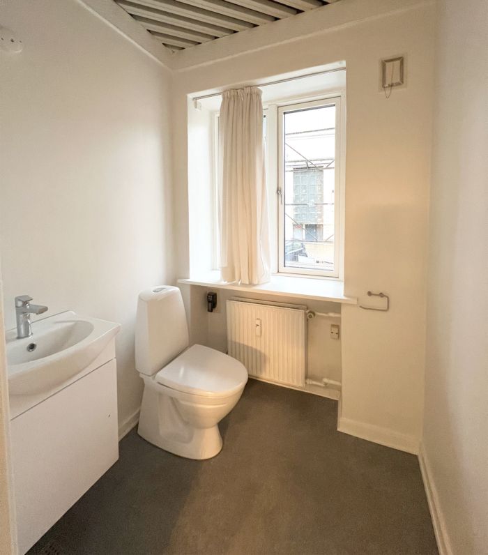 Hvidt badeværelse med hvidt toilet, hvidt vasketøjsskab og hvidt radiator under vinduet med hvid gardin