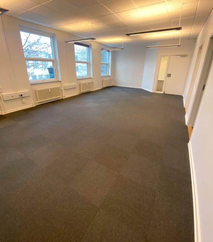 Et tomt kontorlokale med lyse gulve og vægge samt store vinduer med udsigt til en gade med træer og en bygning