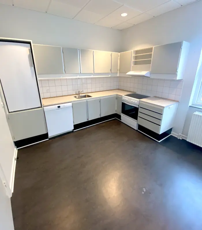 Et moderne køkken med hvide, lavprofilerede skabe, et komfur, en ovn, en vaskemaskine og et køleskab, samt en hvid radiator