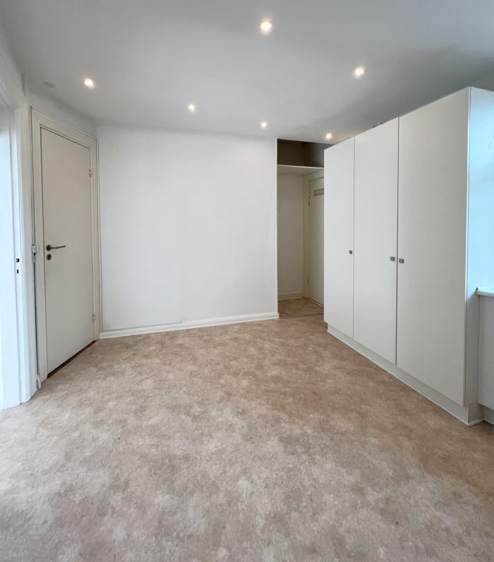 Et rent og moderne lokale med beige gulvbelægning, hvide vægge og indbyggede lysekroner i loftet. Et hvidt skab med