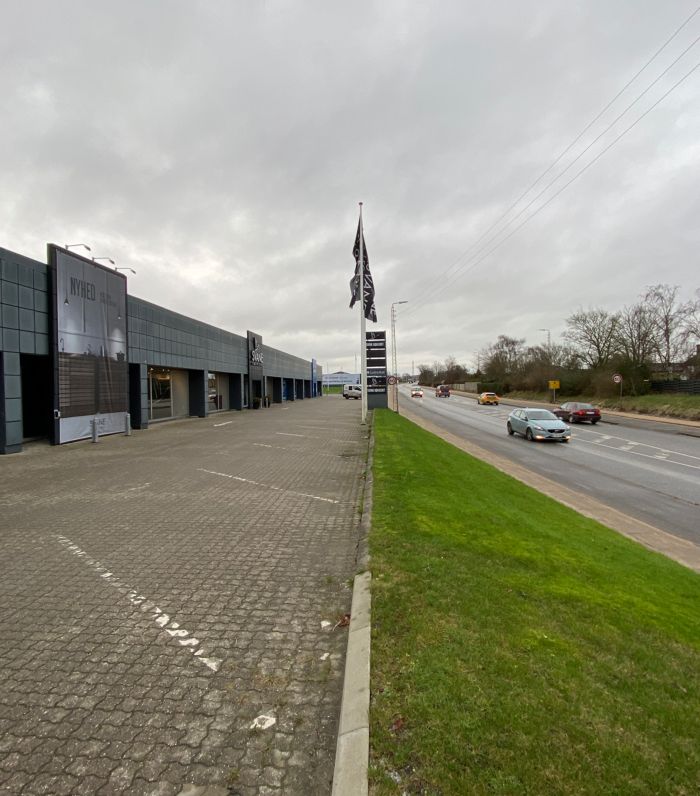 En stor bygning med skiltet Nyhed og et flag i luften langs en motorvej med biler som kører forbi i regnvejr