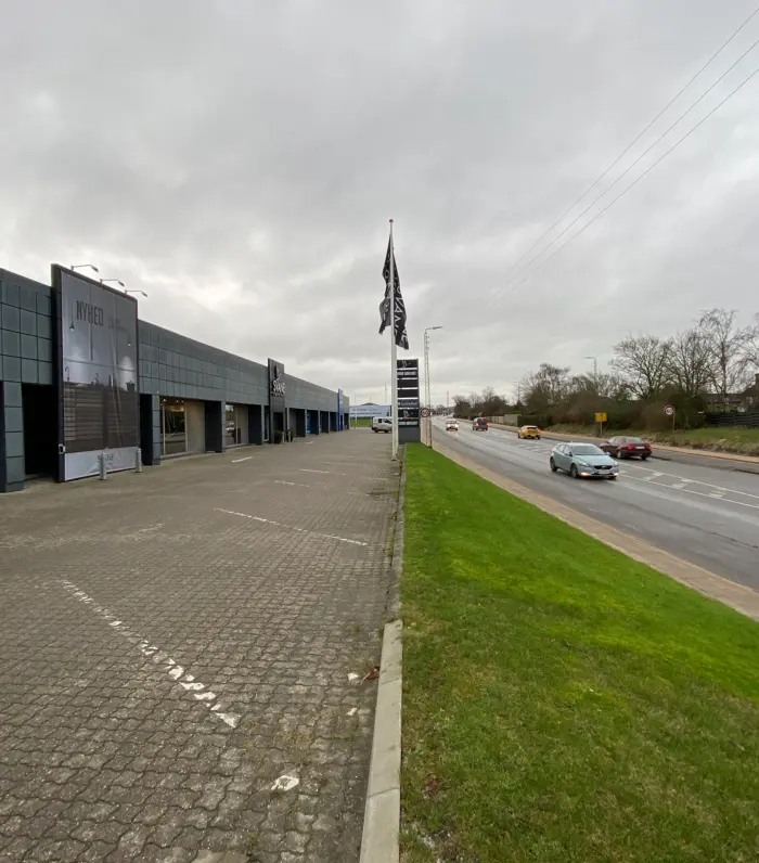 En stor bygning med skiltet Nyhed og et flag i luften langs en motorvej med biler som kører forbi i regnvejr