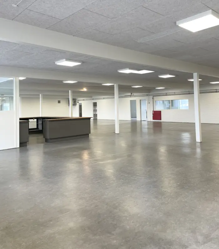 En stor, mørk lagerhal med cementgulv, glasvinduer og hvide vægge samt lyse loftslamper