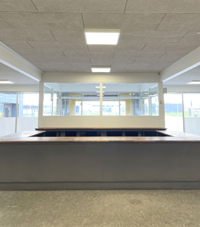 Inde i en stor bygning med et glasvæg og lyse loftsbelysning er der en lang, træfarvet og metalkonstrueret bar med flere