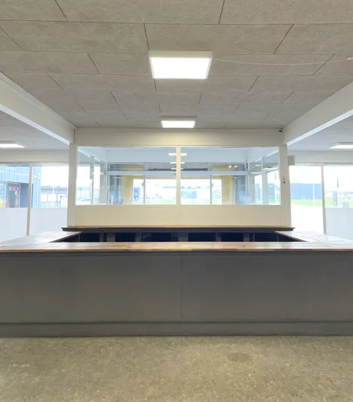 Inde i en stor bygning med et glasvæg og lyse loftsbelysning er der en lang, træfarvet og metalkonstrueret bar med flere