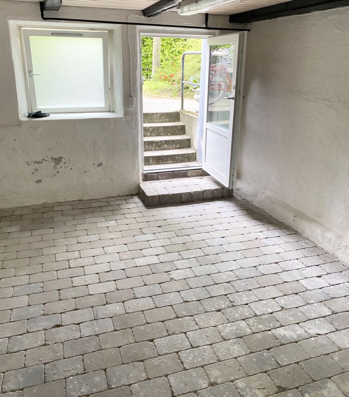 En kælder med tøjlehuse og trapper, der fører ud til gården. Stengulv og hvide vægge med skrald.