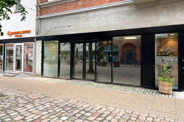 En butik med store glasruder og et plantepotte på fortauet i en gågade med snedækkede stenbrolæg