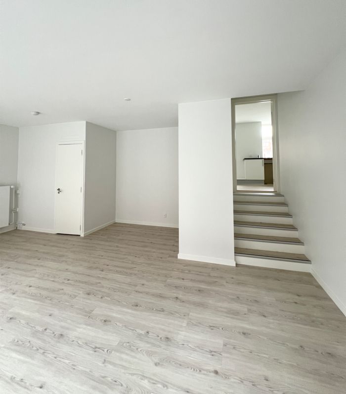 Minimalistisk og nyrenoveret stue med hvide vægge, lyse trægulv og åbne døre til et lyse og luftigt køkken. Stuen er