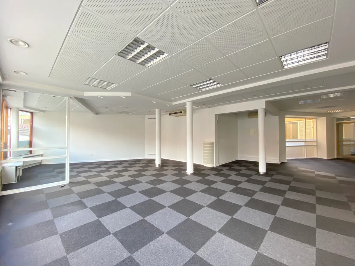 Stort, lyserum med mønstret gulv og hvide vægge og loft med lysskilte. To søjler og klimaanlæg er installeret i rummet. Der
