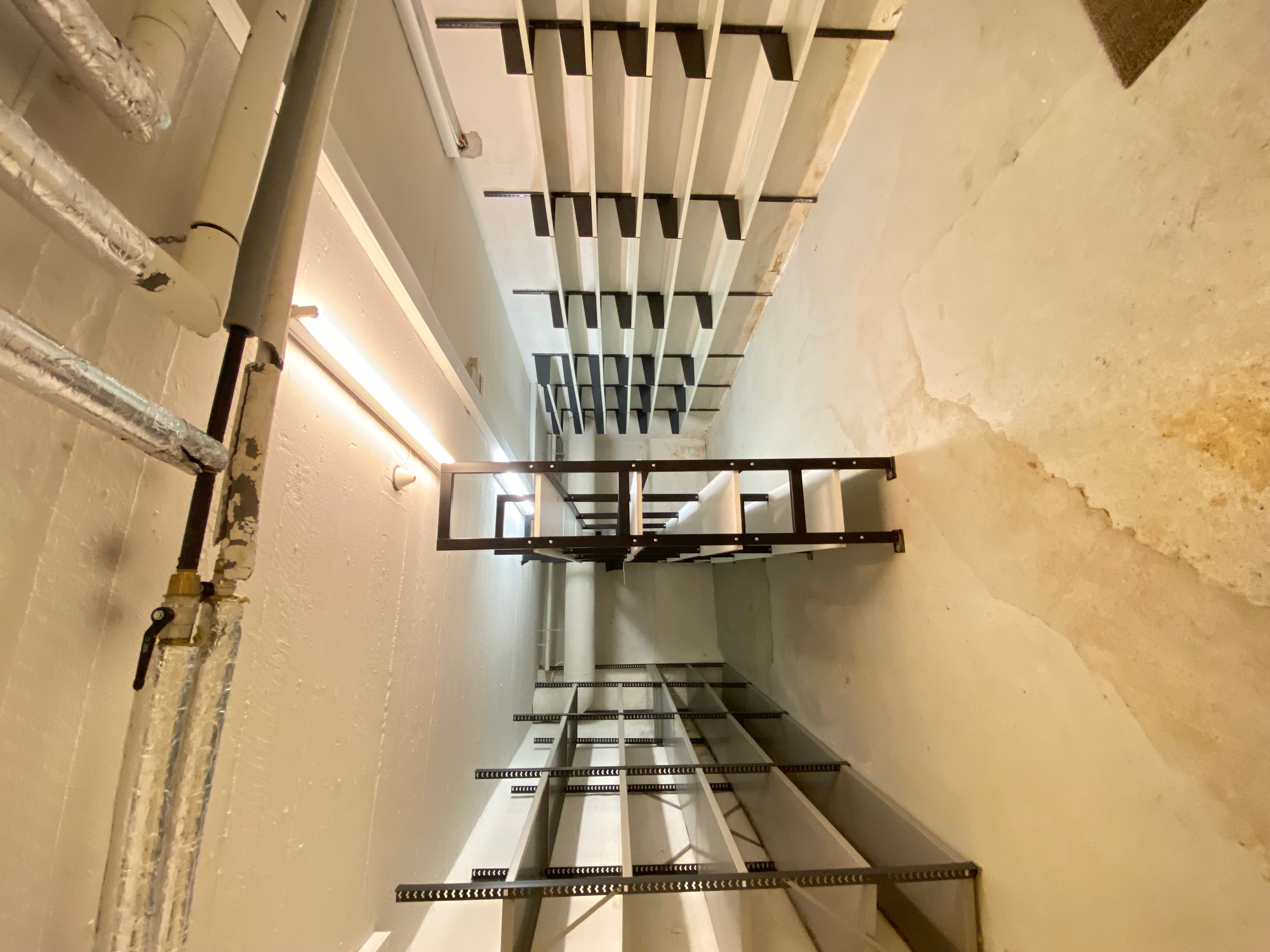 Stålreoler i et rum med højt gulv og hvide vægge med fugtspor og metalrør i siderne