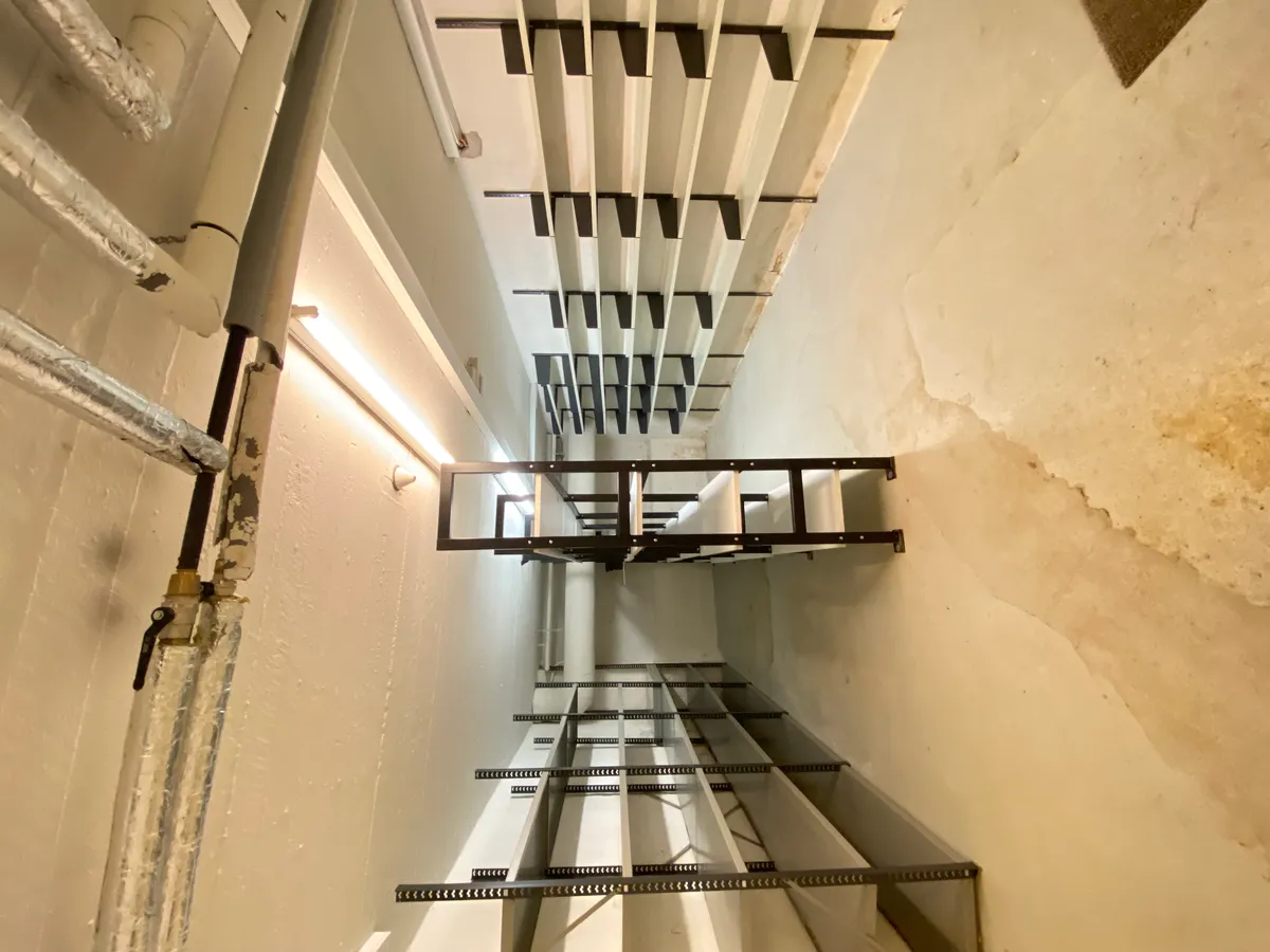 Stålreoler i et rum med højt gulv og hvide vægge med fugtspor og metalrør i siderne