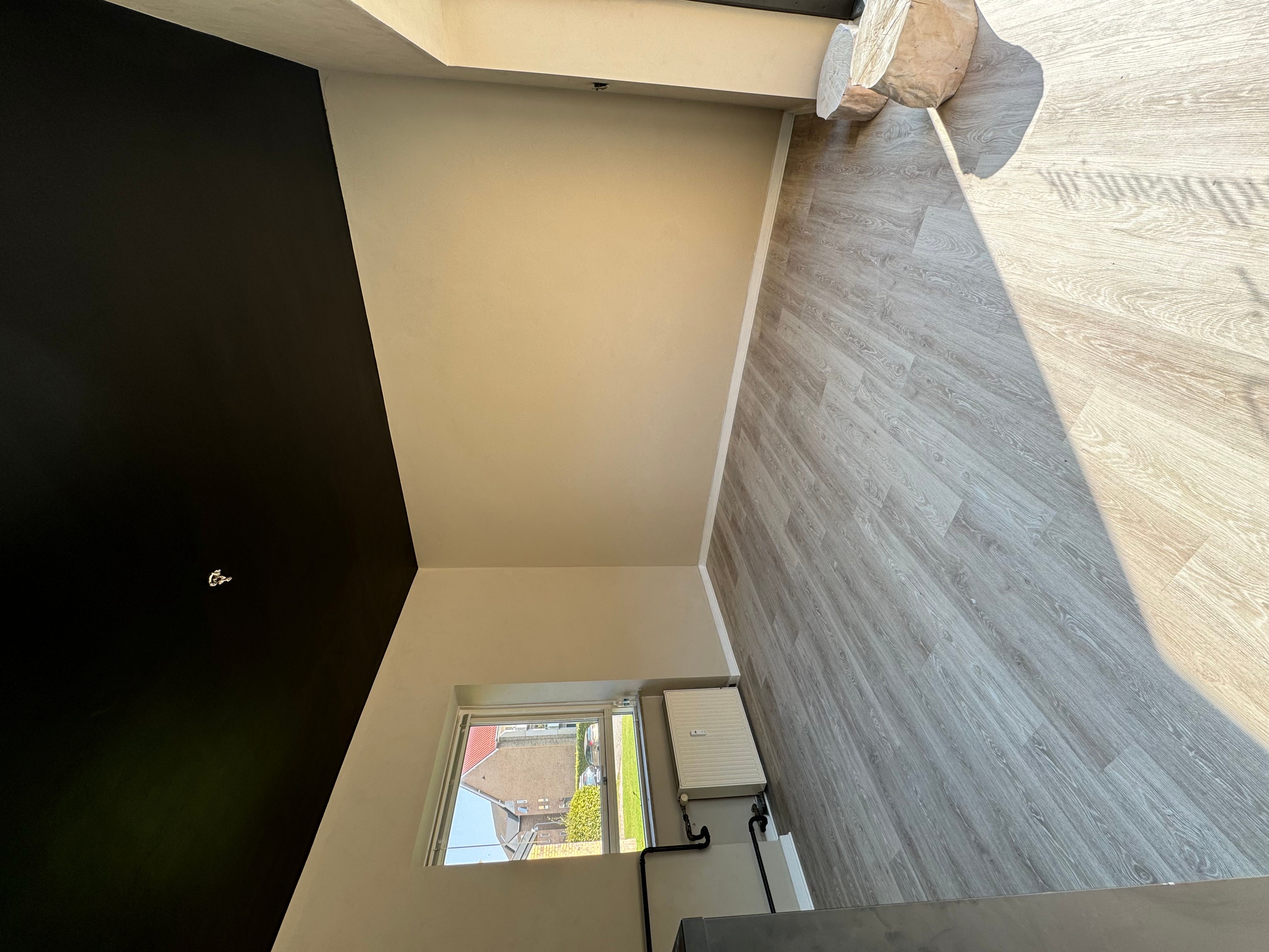 En mørk væg og en lys væg mødes med et hjørne. Der er et vindue med en byudsigt. Der er et træstykke og et radiator på