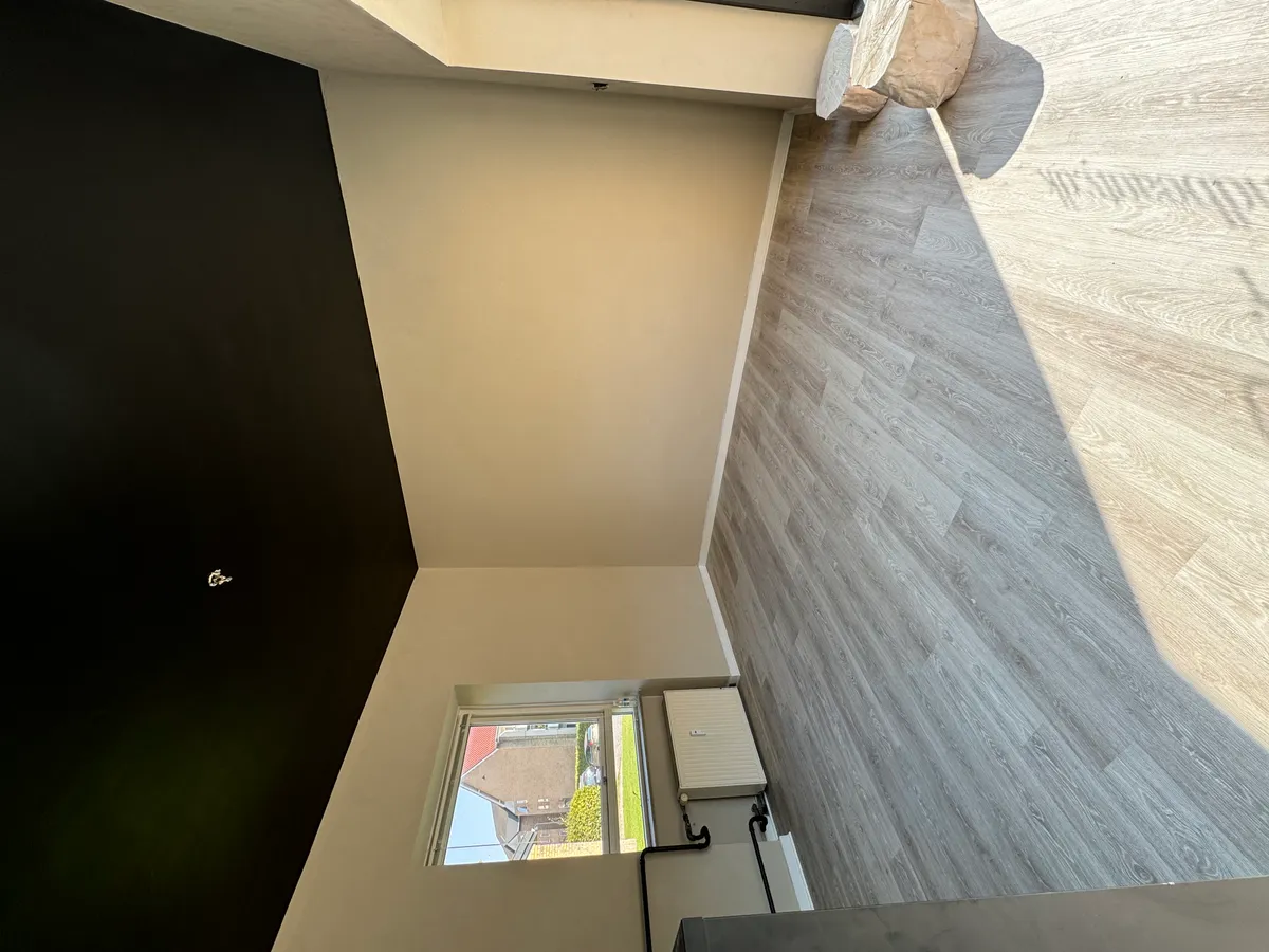 En mørk væg og en lys væg mødes med et hjørne. Der er et vindue med en byudsigt. Der er et træstykke og et radiator på