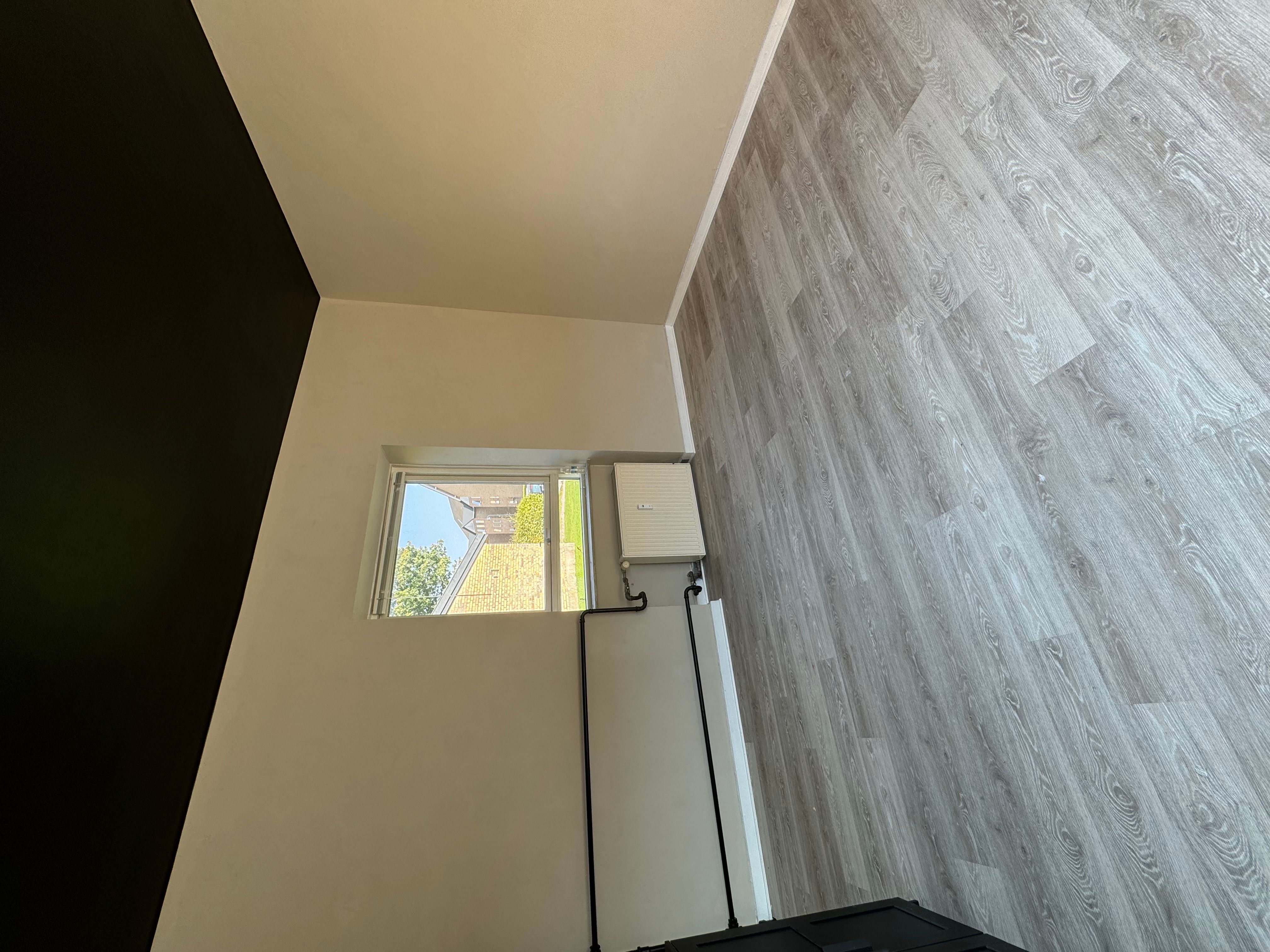 En stor, hvid stue med vindue, hvor der er installeret et varmepumpeanlæg. Der er hårdt trægulv og en sort væg i baggrunden.