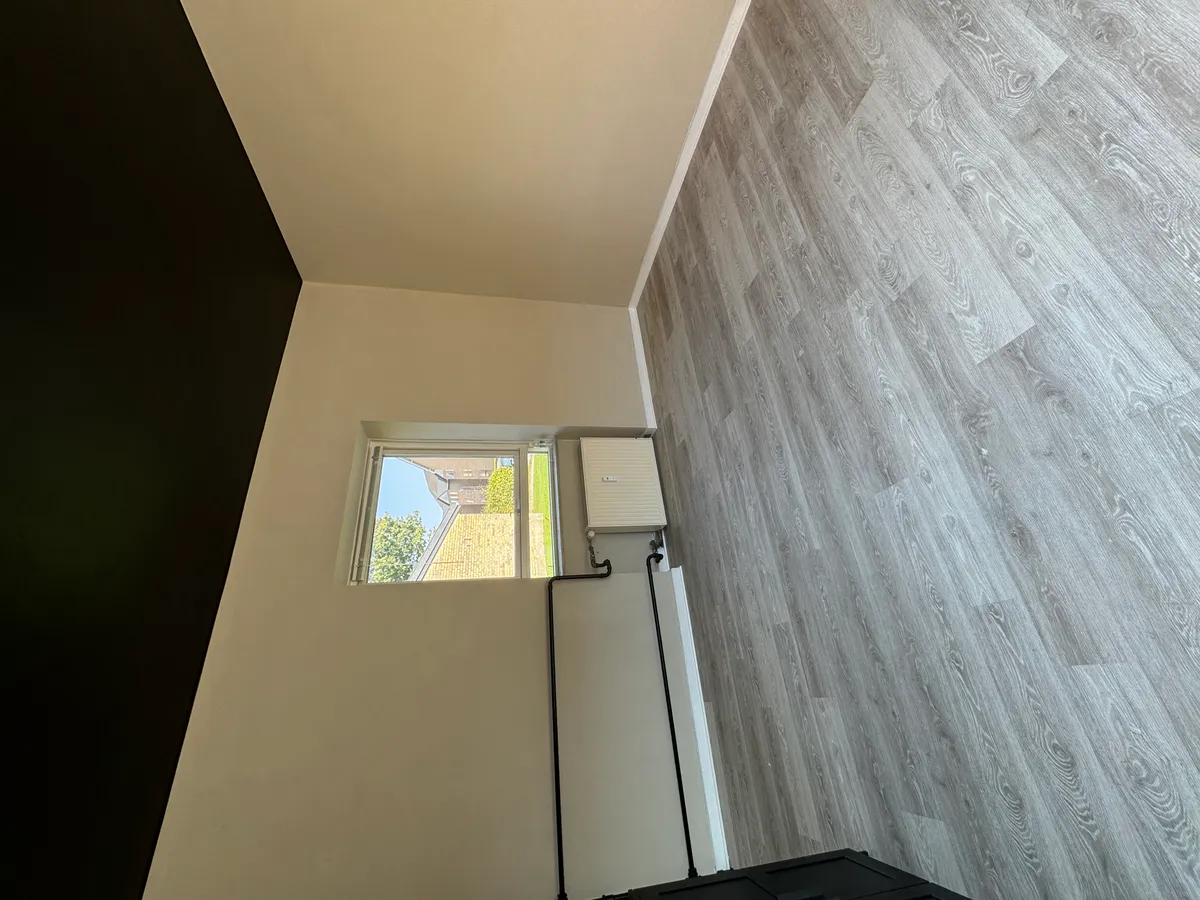 En stor, hvid stue med vindue, hvor der er installeret et varmepumpeanlæg. Der er hårdt trægulv og en sort væg i baggrunden.