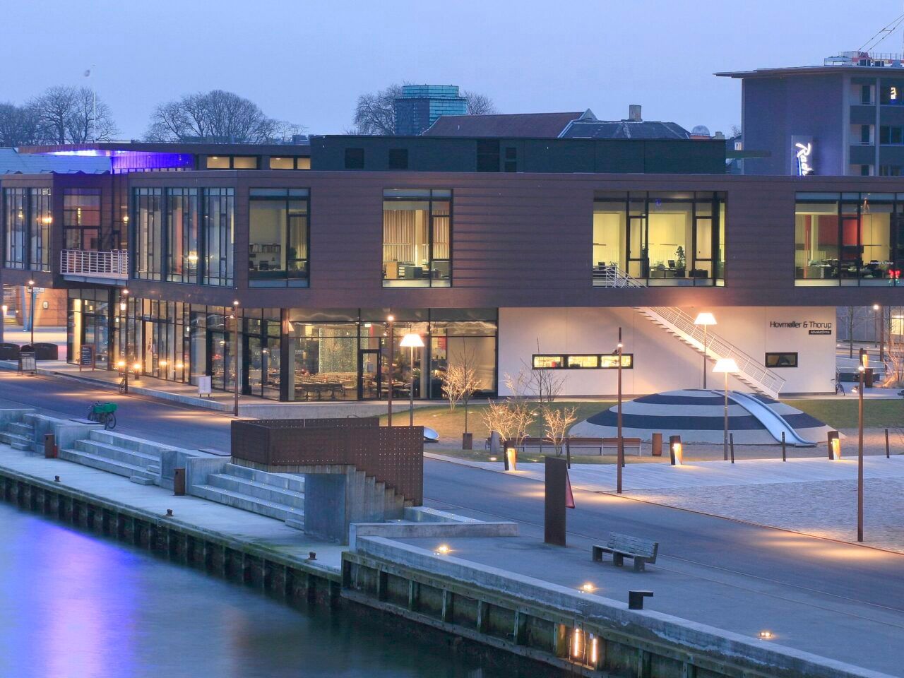 En moderne bygning med glasvinduer og terrasse i centrum med søudsigt, og søen med lyse søvogne og bygninger i baggrunden