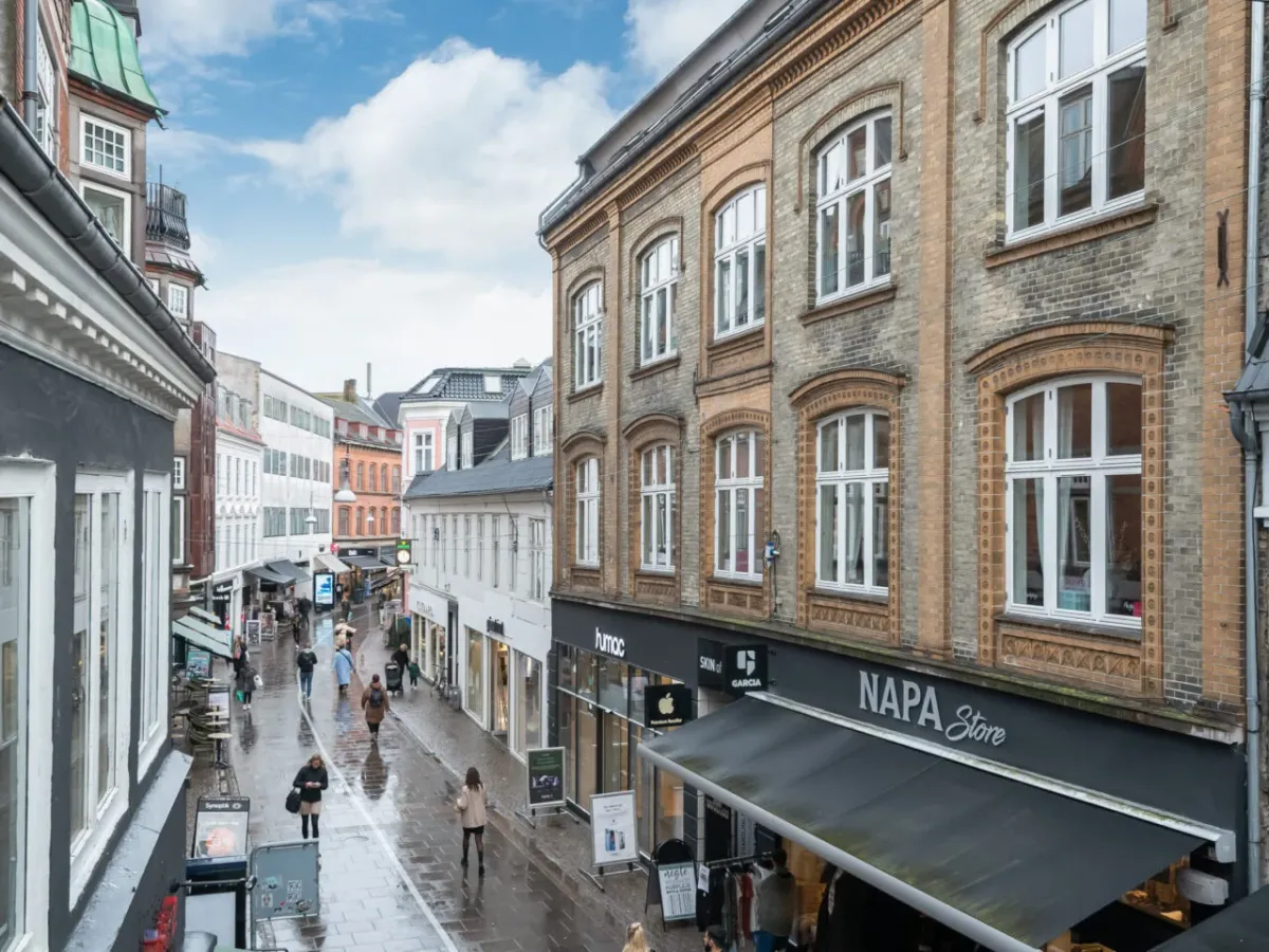 Gågaden Bispensgade i Aalborg med mennesker, forretninger og regnfulde veje, herunder et synligt syntaksmærke og en