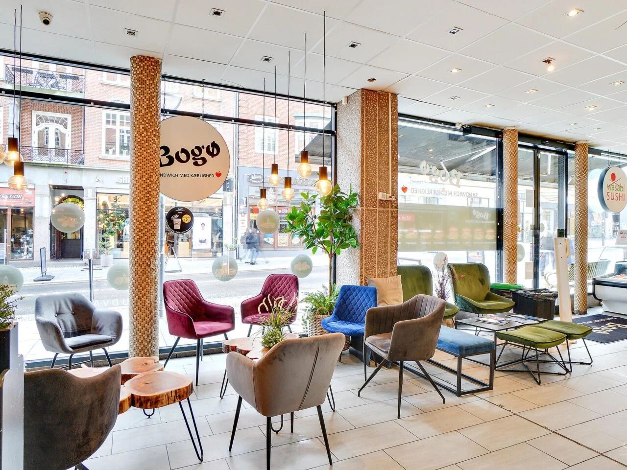 Forretningslokale med store ruder og lyse loftslamper, flere møbler og stole, indretning med farverige stole og runde