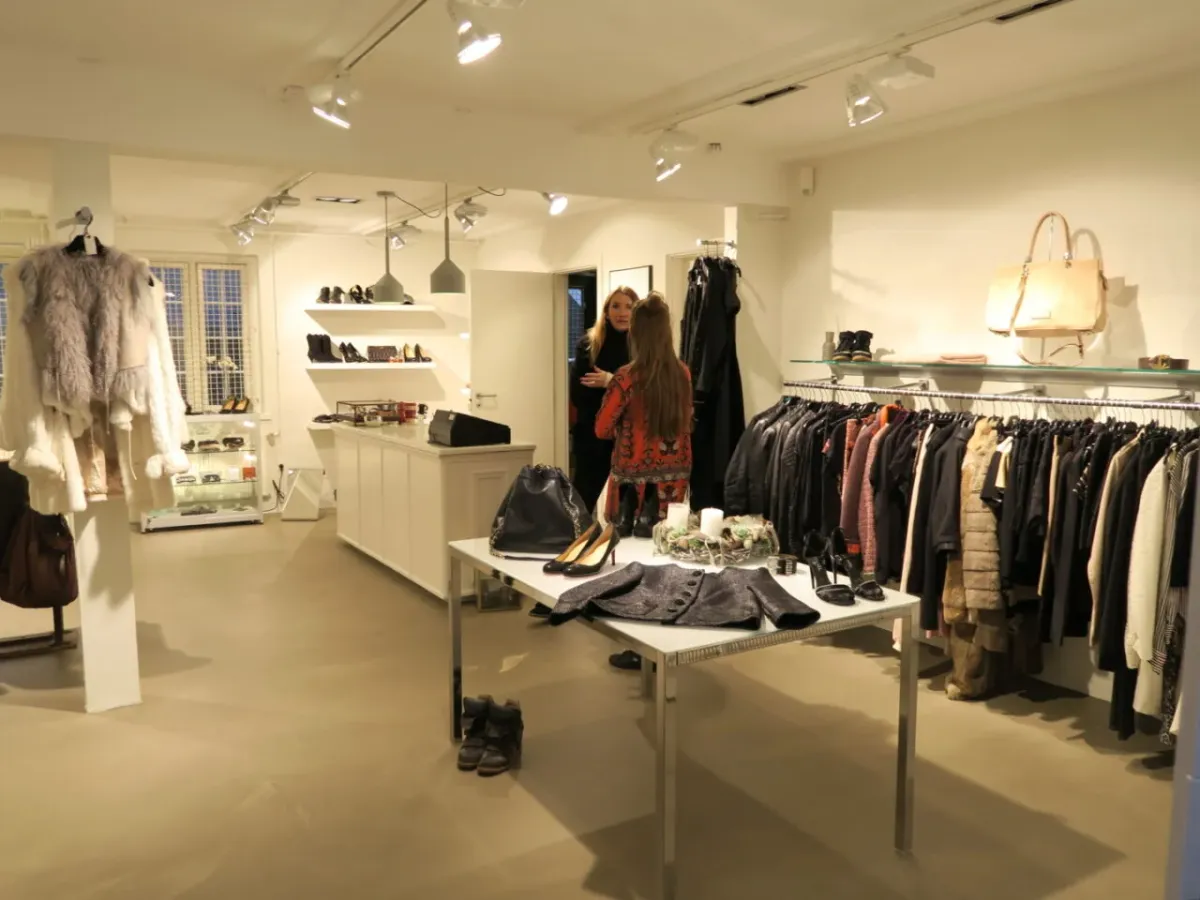 Butik med tøj og sko på gulv og hylder, to kvinder står og ser på tøj og sko, hylder med sko og tøj og flere tøj i klædeskab