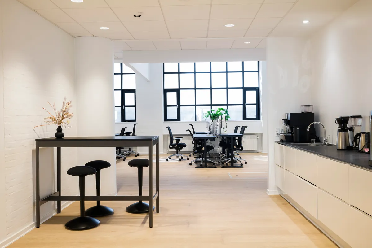 Kontorbord med stole og plante samt skrivebord med kaffemaskine og vandhaner i en hvid indrettet stue