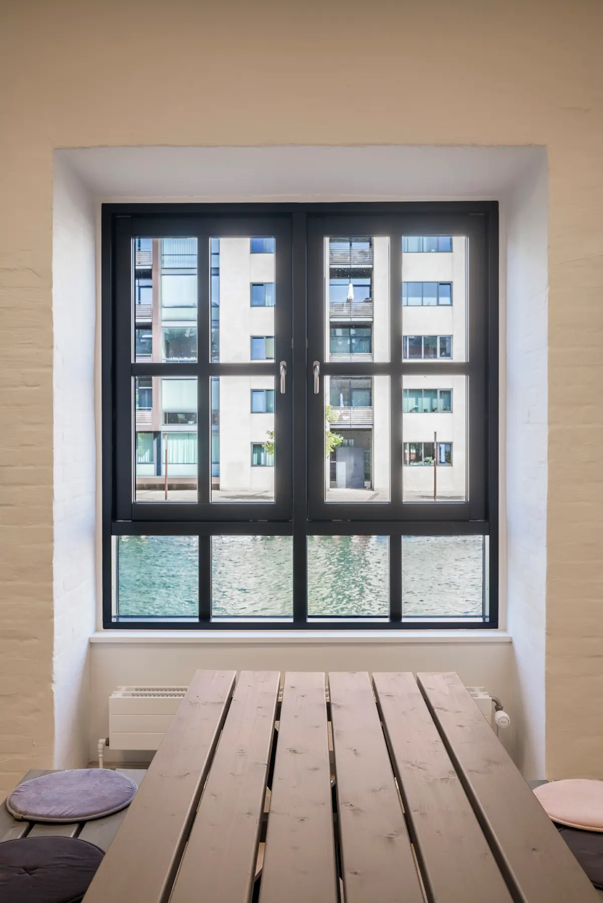 En lukket sort vindue med udsigt til vandet og bygningerne, en træbord og træstole med farvede pudser