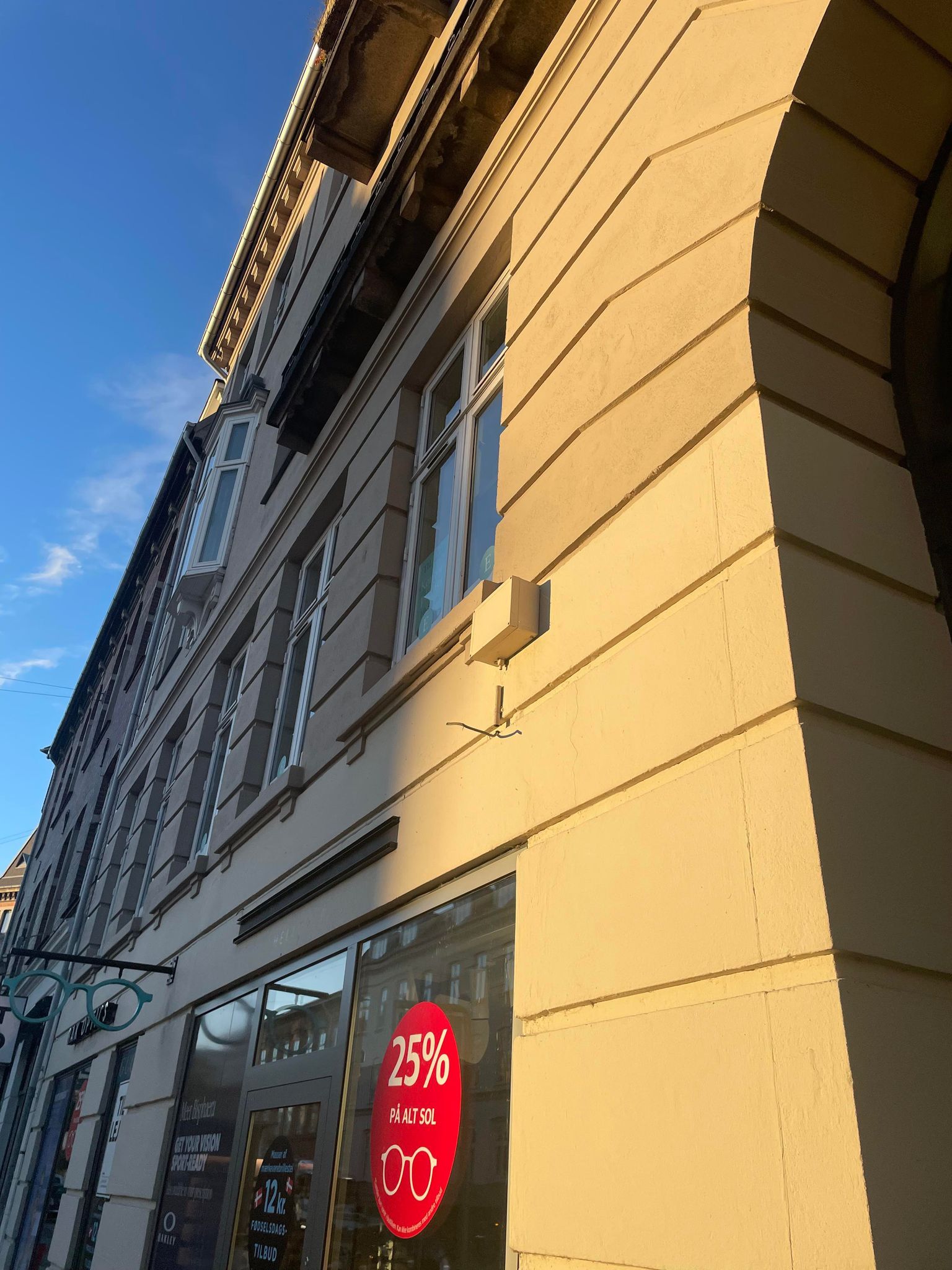 Butik med 25% rabat på alle solbriller. Lysegul væg med møbler og skyggekast. Solbriller hængende udenfor vinduet.