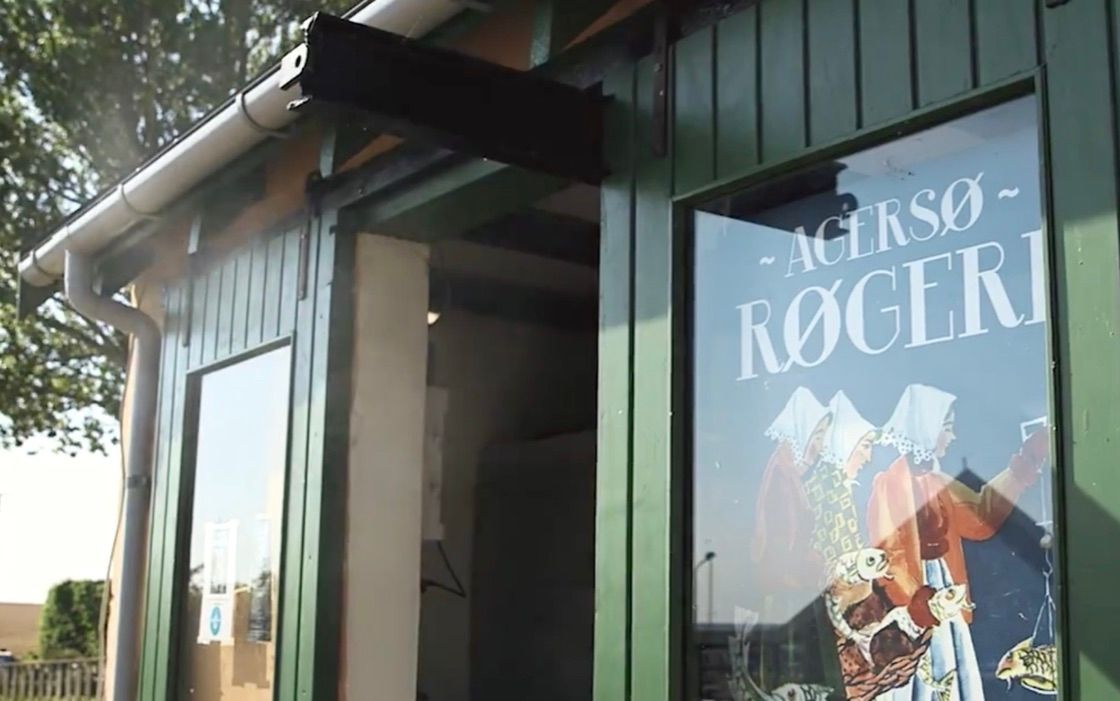 En grøn indgang til en bygning med teksten "Agersø - Rogeri" på vinduet, hvor der er et maleri af to mennesker med fiskekæppe