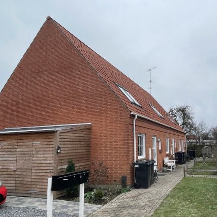 Lejlighedshus i røde mursten med tegltag og hvide ruderuder. Huset har en garage med åbne døre og to skraldespande ved siden