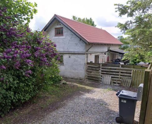 Et hvidt hus med rødt tag og røde vinduesinker på en grusvej med en buske med lilla blomster og en papkasse