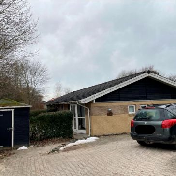 Et lille hus med en stor parkeringsplads foran og en SUV parkeret. Huset har et sort tag og en terrasse med vinduer. Et