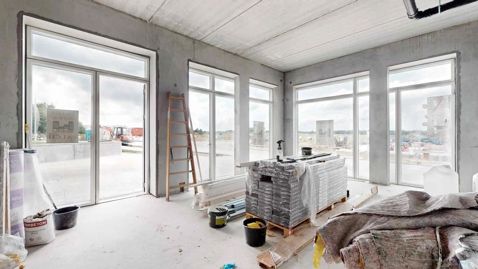 Konstruktionsområde med flere vinduer og glasdøre, hvor der er træstiger, emballage, og byggematerialer samt byggemaskiner