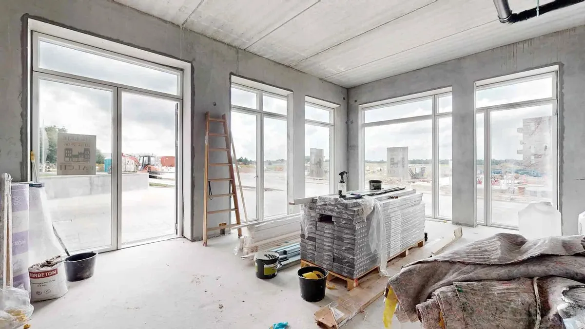 Konstruktionsområde med flere vinduer og glasdøre, hvor der er træstiger, emballage, og byggematerialer samt byggemaskiner
