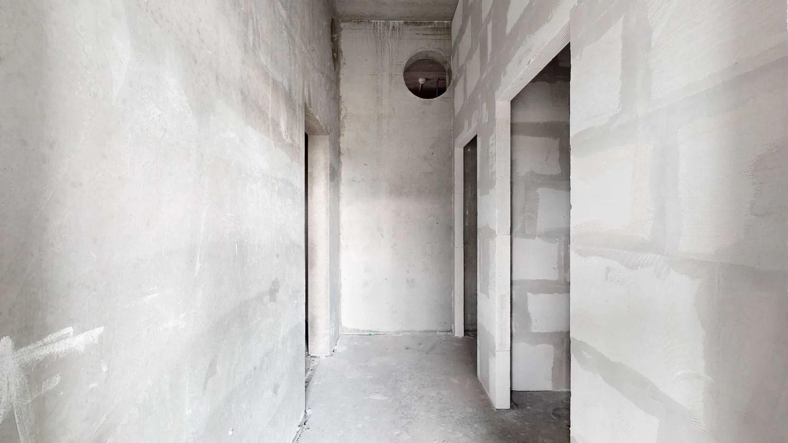 En stor nybygget korridor med hvide vægge og cementgulv, to døre og et rundt hul i loftet