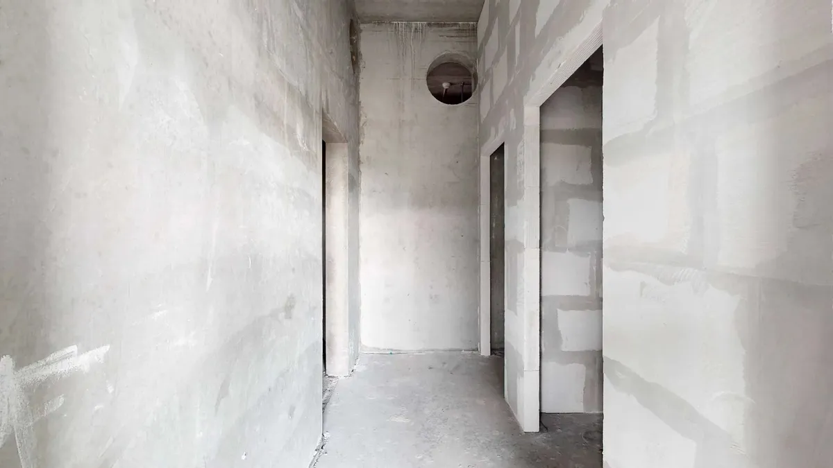 En stor nybygget korridor med hvide vægge og cementgulv, to døre og et rundt hul i loftet