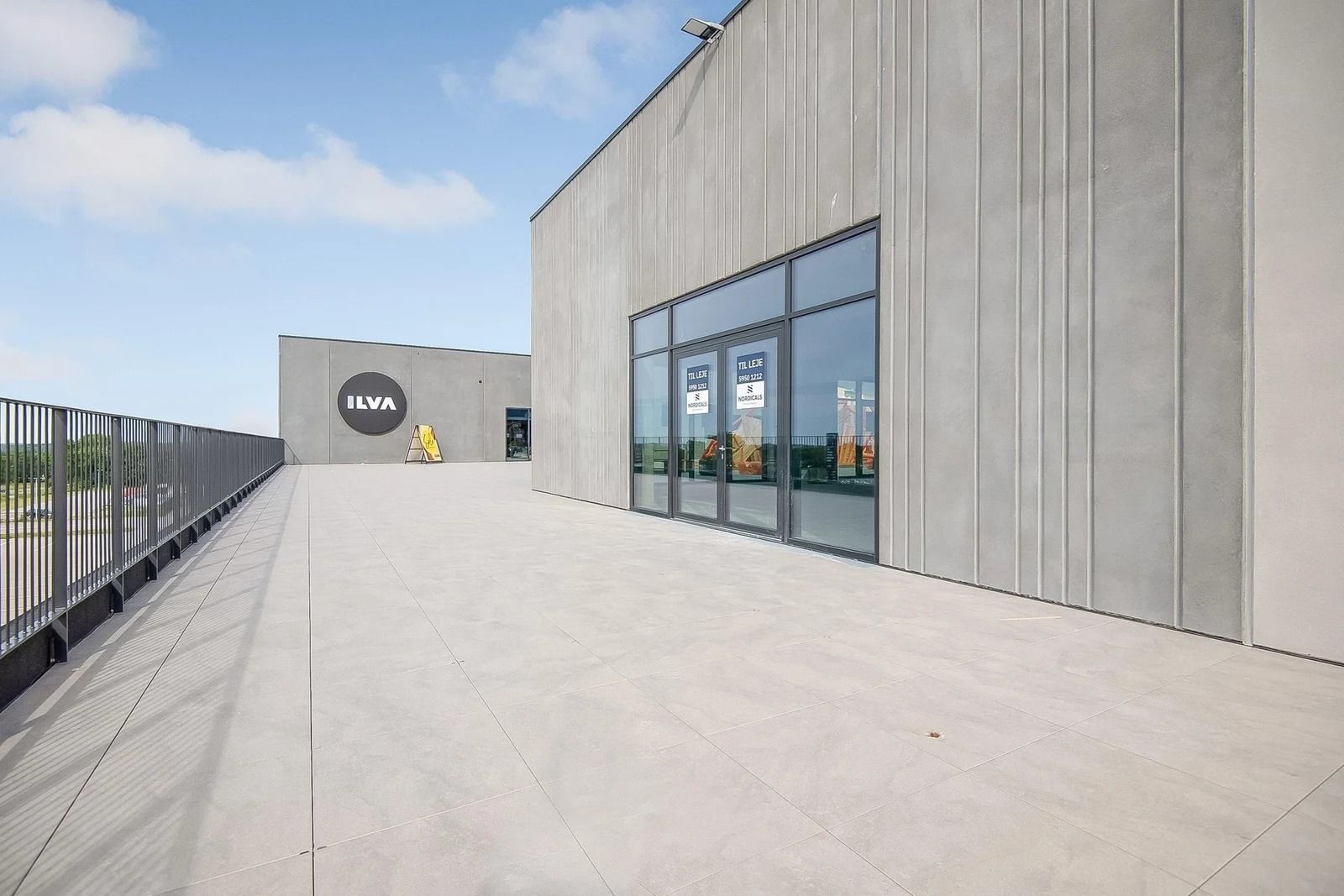 Lejlighedsbygning med store glasruder og logoet ILVE. Der er parkeringsplads med et skilt og en metalhegn. Bygningen er i