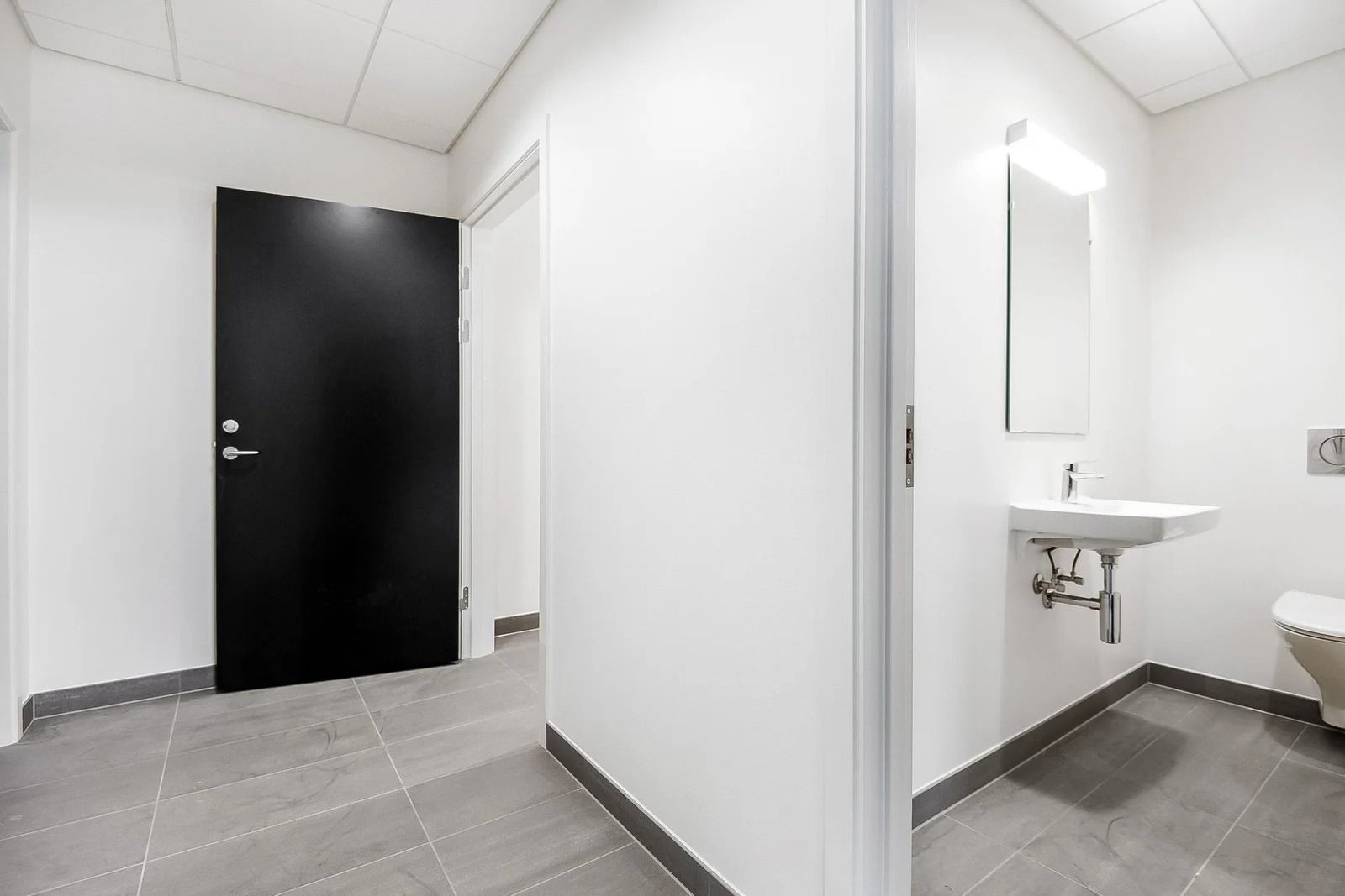 En minimalistisk og stilfuldt designet toiletrum med hvide vægge, gulv og loft, en sort dør, en spejl og en hvid vask samt