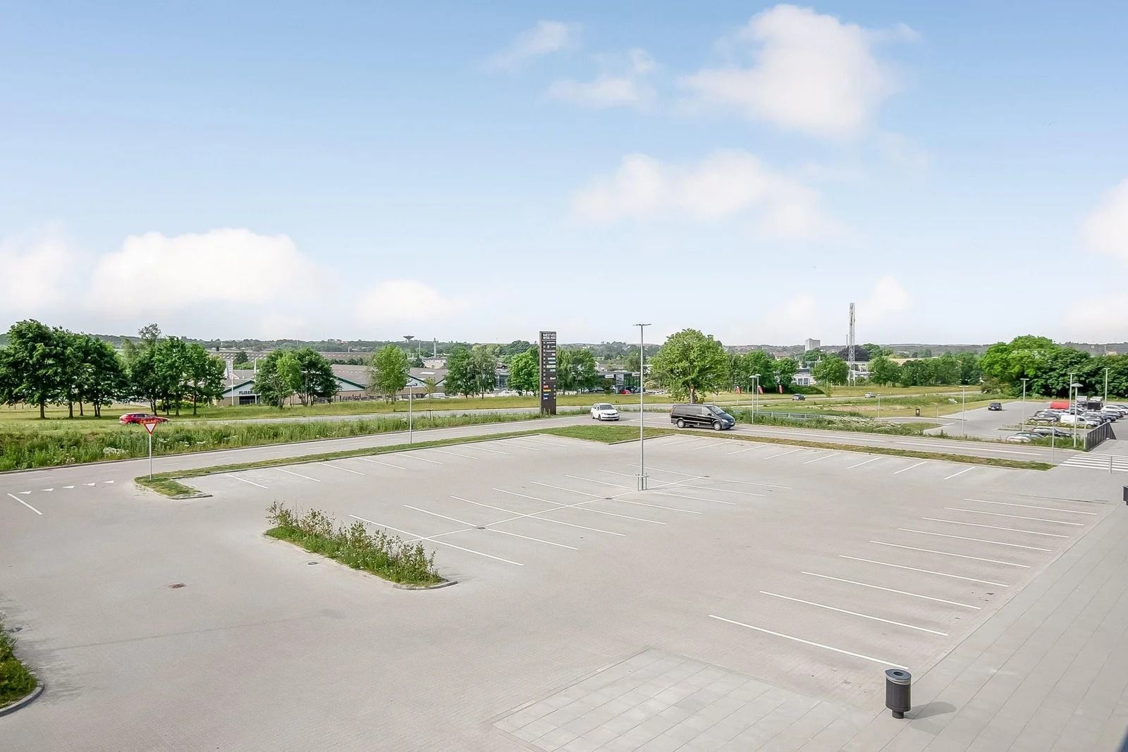 En stor parkeringsplads med to biler og et drejeskilt og flere grønne buske og træer langs siderne og flere biler parkeret