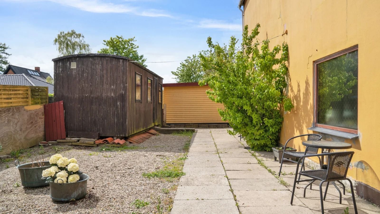 En lille gårdhave med et gammelt hyttehus og en gul stenmur, der er dækket med grønne planter og blomster