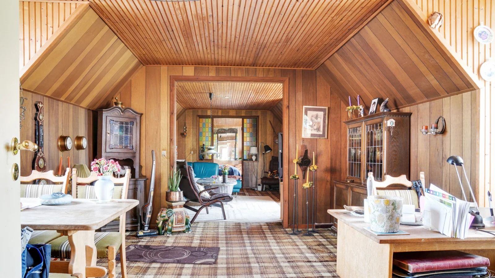 En værelse med lyse, træpanelerede vægge og loft, møbler og tavler, samt en lampe og en sofa i midten af rummet