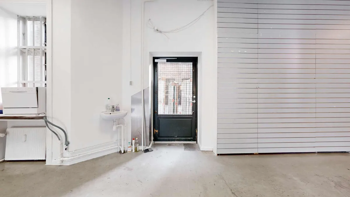 En stor, lys og ren stue med hvide vægge, lysegul gulv og en hvid dør i midten.