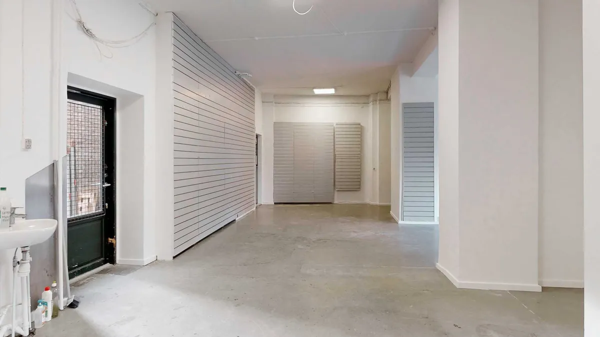 En stor, lysegrå, tom lejlighed med to store vinduer og døre i baggrunden, samt en sink med vandhane og en åben dør til en