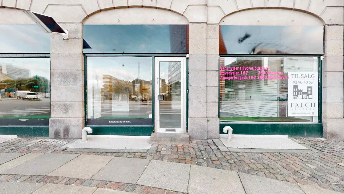 Bygning med to vinduer og en dør. Butiksvinduet er lukket og har skilt med tekst og nummer. Bag vinduet ses en bygning og