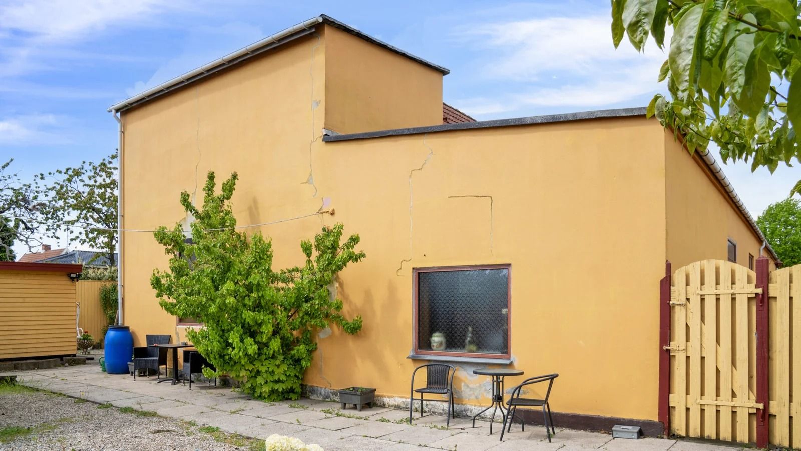 Gult hus med gitterdør, udsigt til et lille terrassebord med stole, og en plante i forgrunden