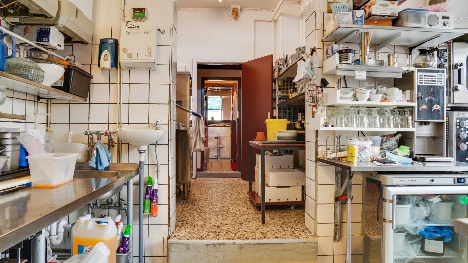 Indkøbskurv og flasker med rengøringsmidler på et køkkenbord med et vaskebækken og flasker med rengøringsmidler på hylder i