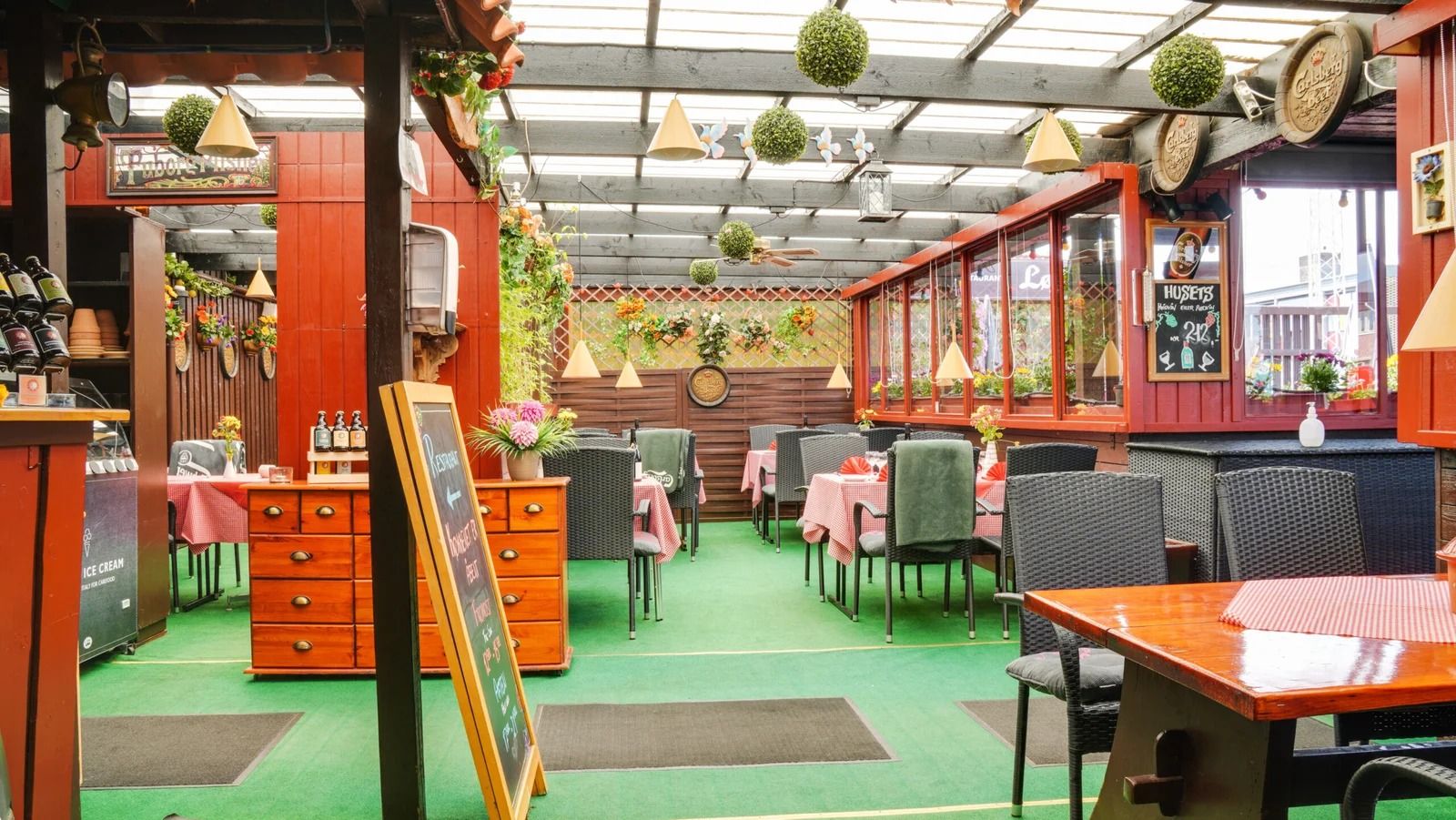 Lysthus med træborde og stole, indretning med bordplade, og hængende lamper i lyse farver og grønne planter