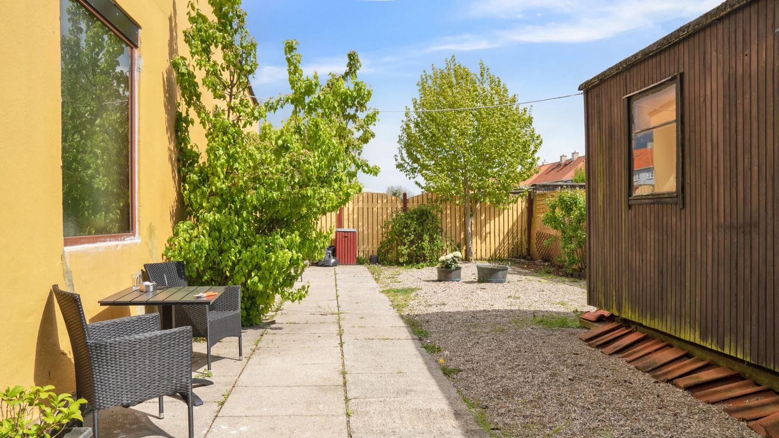 En terrasse med bord og stole, en blomsterpotte og et rødt skur i baggrunden, samt en gågade og en høj gul mur