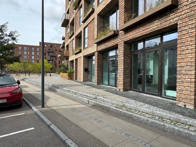 En rød bil er parkeret på siden af en gågade, hvor der er et stort røde murstensbygning med glasruder