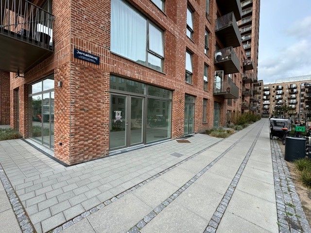 En moderne bygning med glasvinduer og en højtliggende balkon. En smal og lang vej fører forbi bygningen. Foran bygningen er